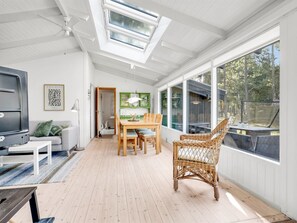 Ein geräumiges Zimmer mit Holzboden, einem Essbereich mit Tisch und Stühlen, einem Korbstuhl, einem Fernseher und einem großen Fenster mit Blick auf Bäume.