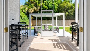 Eine überdachte Terrasse mit Deckenventilator, Tisch, Stühlen und Blick auf einen Garten mit Holzlattenzaun und Grünpflanzen.