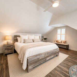 Une chambre à coucher avec un cadre de lit en bois, une lampe de chevet, un banc avec des coussins, une fenêtre et un ventilateur de plafond.