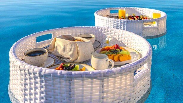 Un'area piscina con sedie in vimini, cibo e bevande, acqua cristallina e vista sullo sfondo del mare.