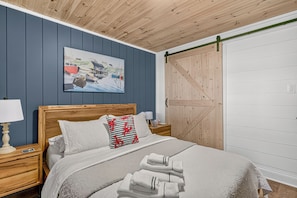 A bedroom with a wooden headboard, a painting of a harbor scene, a sliding barn door, and a neatly made bed with towels. - Hot Tub | Sauna | Oceanfront |  Fall Colours | near Peggy’s Cove & Halifax (Prospect Village)