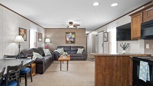 Et moderne køkken med en central ø, et spiseområde med bord og stole, en stue med sofa og loftventilator samt en køkkenbordplade med hårde hvidevarer.