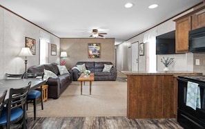 A modern kitchen with a central island, a dining area with a table and chairs, a living room with a sofa and a ceiling fan, and a kitchen counter with appliances.