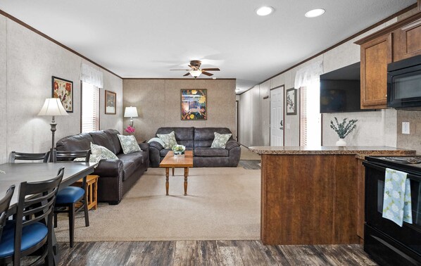 A modern kitchen with a central island, a dining area with a table and chairs, a living room with a sofa and a ceiling fan, and a kitchen counter with appliances.
