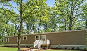 A single-story mobile home with a front porch, a small deck, and a front yard with a tree and flower beds.