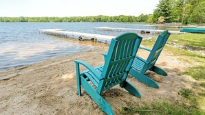 Uitzicht op een meer met een steiger, een rij groene Adirondack-stoelen en een zandstrand.