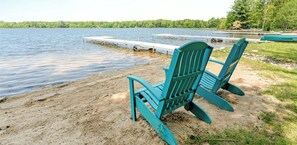 Blick auf einen See mit Steg, einer Reihe von grünen Adirondack-Stühlen und einem Sandstrand.