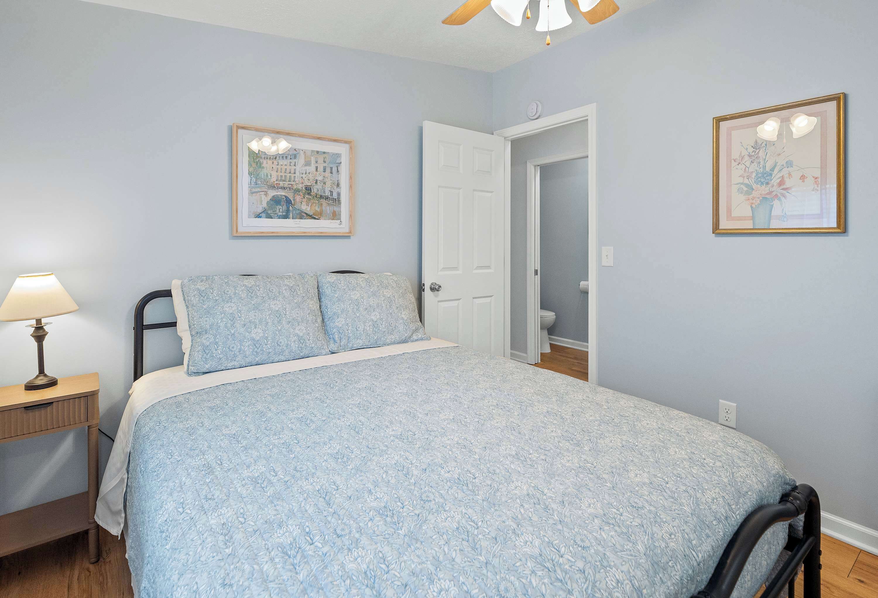 A bedroom with a bed, a nightstand, a lamp, a door, and two framed pictures on the wall.