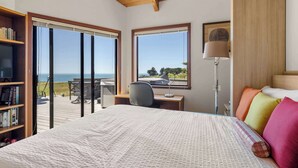Ein Schlafzimmer mit einem großen Bett, einem Schreibtisch, einem Stuhl, einem Bücherregal und Blick auf das Meer durch große Fenster.