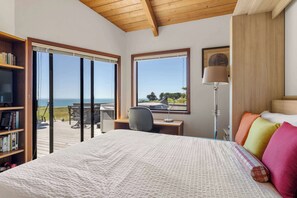 Ein Schlafzimmer mit einem großen Bett, einem Schreibtisch, einem Stuhl, einem Bücherregal und Blick auf das Meer durch große Fenster.