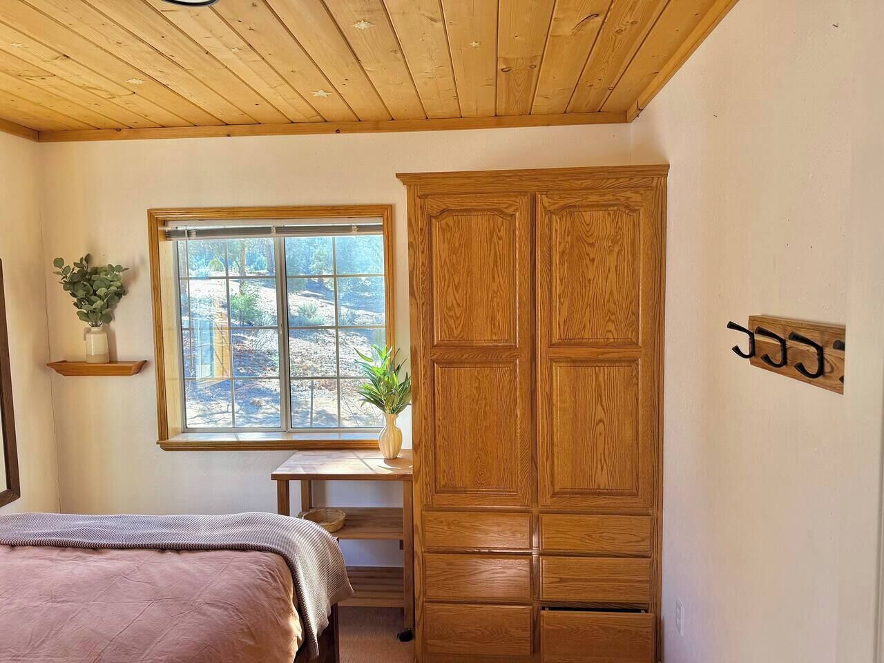 A bedroom with a wooden wardrobe, a bed, a window with a view, and a small desk with a plant.