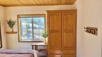 A bedroom with a wooden wardrobe, a bed, a window with a view, and a small desk with a plant.