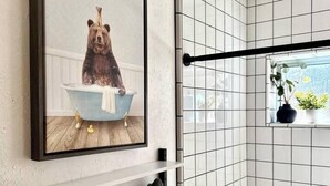 A bathroom with white tiled walls, a framed picture of a bear in a bathtub, a towel rack with a green towel, and a window with a plant.