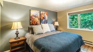 A bedroom with a bed, two bedside lamps, a window with a view of greenery, and two abstract paintings on the wall. - Garvey House Red | Downtown Home with Hot Tub (Red Lodge)