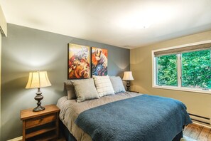 A bedroom with a bed, two bedside lamps, a window with a view of greenery, and two abstract paintings on the wall. - Garvey House Red | Downtown Home with Hot Tub (Red Lodge)