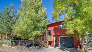 A red two-story house with a front deck, garage, and surrounding trees. - Garvey House Red | Downtown Home with Hot Tub (Red Lodge)