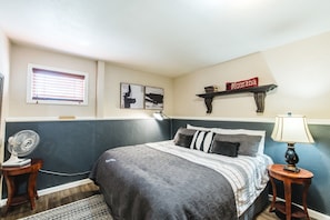 A bedroom with a bed, bedside tables, a wall-mounted shelf, a window with blinds, and a fan.
