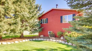 A red building with a stone border, a large tree, and a small bush. - Garvey House Red | Downtown Home with Hot Tub (Red Lodge)