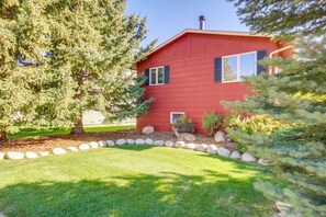 A red building with a stone border, a large tree, and a small bush. - Garvey House Red | Downtown Home with Hot Tub (Red Lodge)