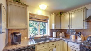 A kitchen with wooden cabinets, a built-in oven, a coffee maker, and a window with a view of trees. - Garvey House Red | Downtown Home with Hot Tub (Red Lodge)