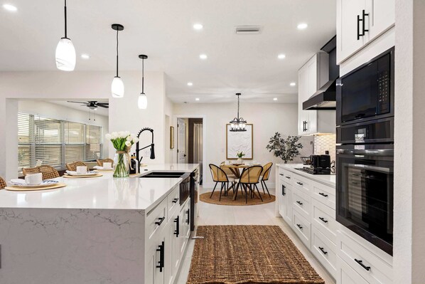 Una cocina moderna con armarios blancos, una isla de mármol, iluminación colgante y un comedor con una mesa redonda y sillas.