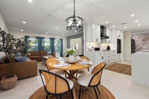 Una sala moderna con sofá, mesa de comedor y sillas, una cocina con armarios blancos, y un ventanal con cortinas.