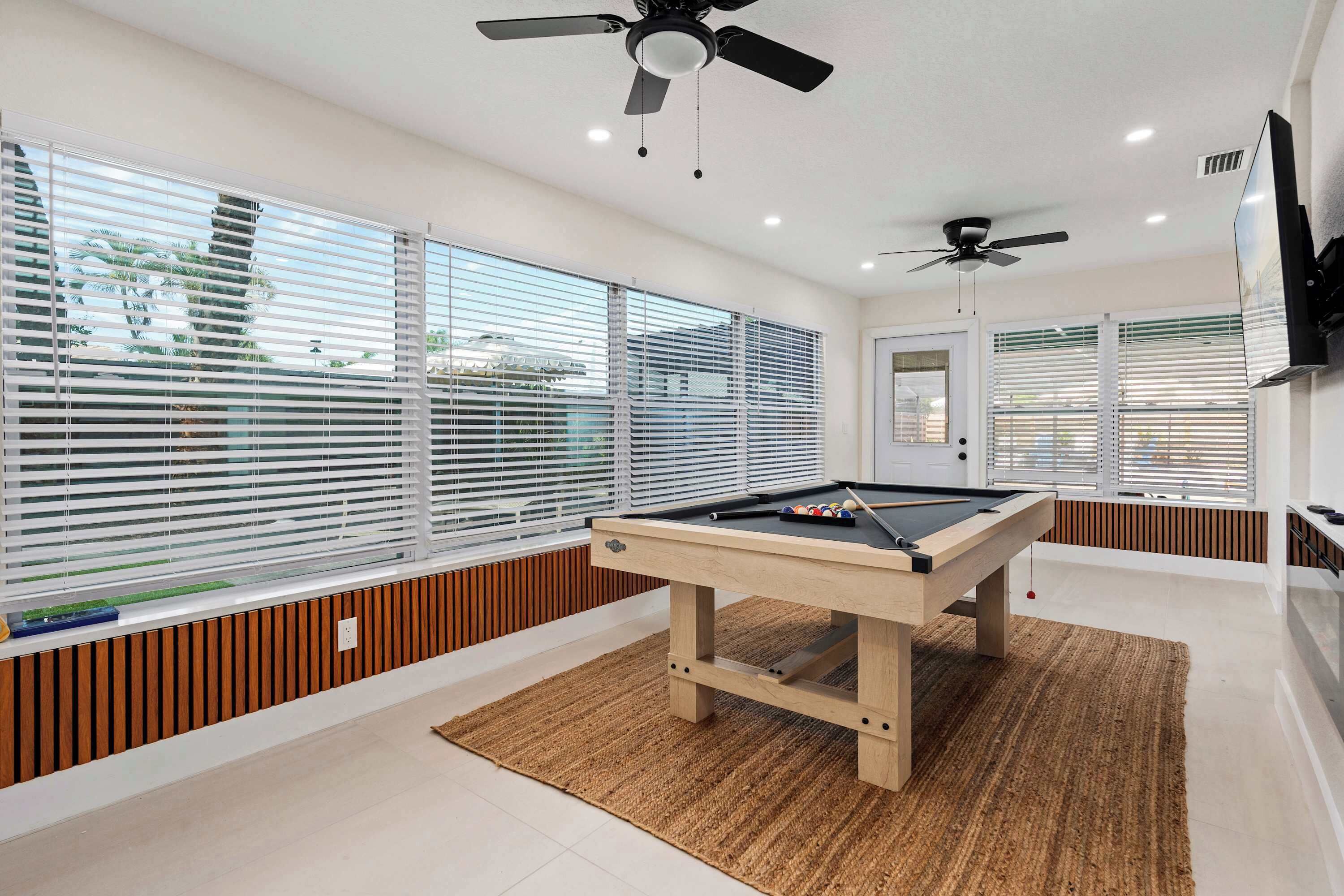 A pool table in a room with large windows, ceiling fans, and a television mounted on the wall.