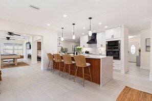 Una cocina moderna con una isla central, taburetes de bar, iluminación colgante y una zona de comedor con mesa de billar.