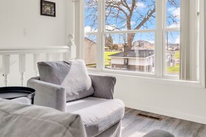 A room with a grey armchair, a small table, and a window with a view of houses and trees.