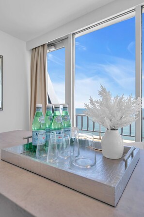 Une chambre d’hôtel avec une grande fenêtre, un plateau avec des bouteilles et des verres, et un vase blanc contenant des fleurs blanches.