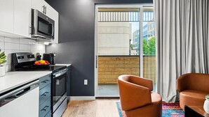 Een moderne keuken met een magnetron, fornuis en vaatwasser, een schuifpui naar een balkon, en een bruine leren fauteuil.