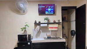 Een kleine keuken met een magnetron, spoelbak en kastjes, een ventilator en een tv aan de muur.