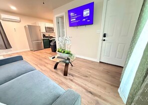 Ein modernes Wohnzimmer mit einem grauen Sofa, einem Glastisch mit Pflanzen, einem an der Wand befestigten Flachbildfernseher und einer Küche im Hintergrund.