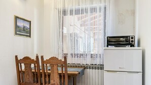 Una pequeña cocina con un refrigerador blanco, una mesa de comedor de madera con sillas y una ventana con cortina translúcida.