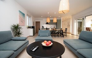 Ein modernes Wohnzimmer mit einer Sofagarnitur, einem Couchtisch mit Früchten und Fernbedienungen sowie einer Küche im Hintergrund.