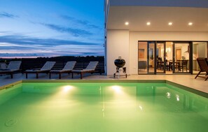 A modern outdoor pool area with lounge chairs, a barbecue grill, and a glass door leading to a dining area. - Gorgeous home in Medulin (Medulin)