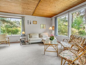 Ein gemütliches Zimmer mit Holzbalkendecke, einer Couch, einem Tisch mit einer Vase, Korbstühlen und großen Fenstern mit Vorhängen.
