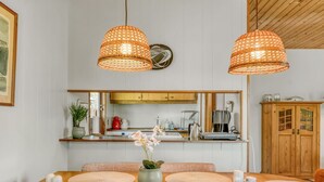 Een eethoek met een houten tafel, geweven hanglampen, een vaas met bloemen en een keuken op de achtergrond.