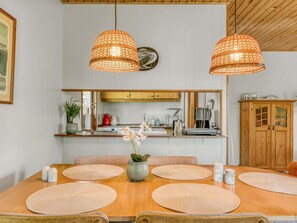 Ein Essbereich mit einem Holztisch, gewebten Pendelleuchten, einer Vase mit Blumen und einer Küche im Hintergrund.