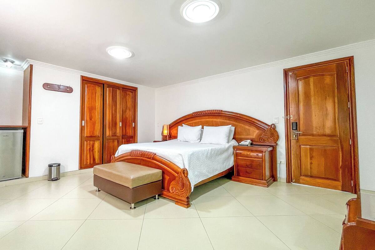 A bedroom with a wooden bed, a nightstand, a bench, a wardrobe, and a small refrigerator.