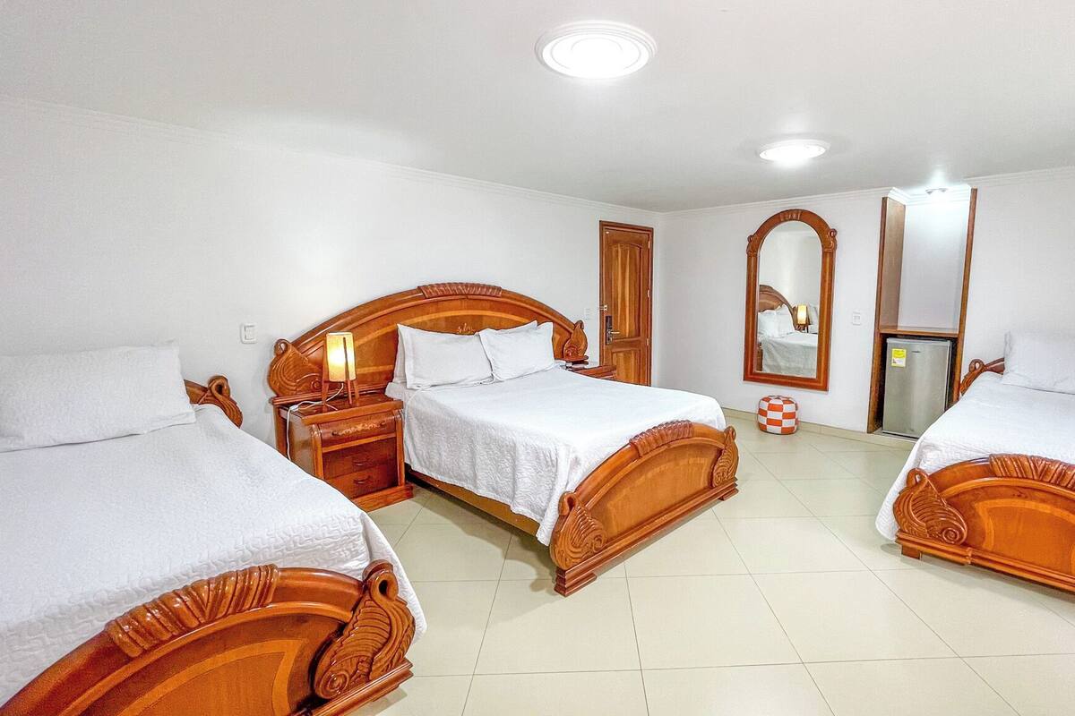 A hotel room with three wooden beds, a small refrigerator, a bedside table with a lamp, and a mirror on the wall.