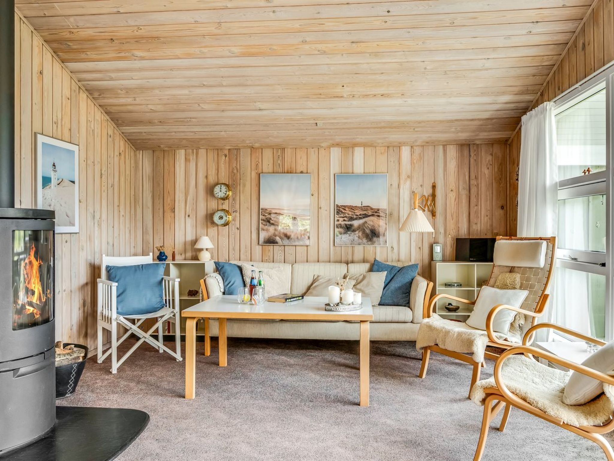 A cozy cabin interior with a fireplace, wooden walls, a sofa, chairs, and artwork.