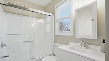 Un baño con cabina de ducha de vidrio, inodoro, lavamanos con espejo y ventana.