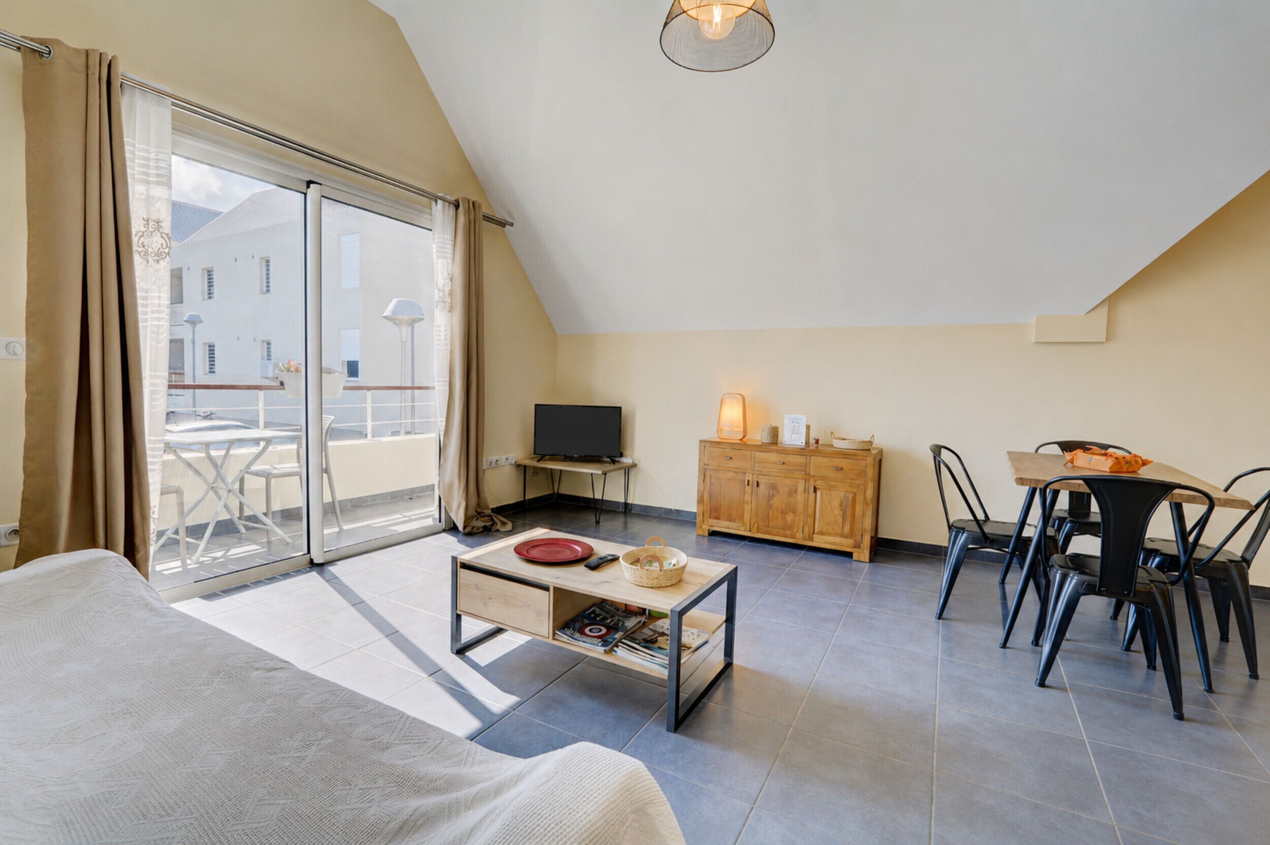 A room with a bed, a dining table with chairs, a TV, a wooden cabinet, and a balcony with a table and chairs.