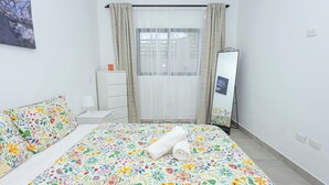 Ein Schlafzimmer mit floralem Bettbezug, einer weißen Kommode, einem Spiegel und einem Fenster mit Vorhängen.