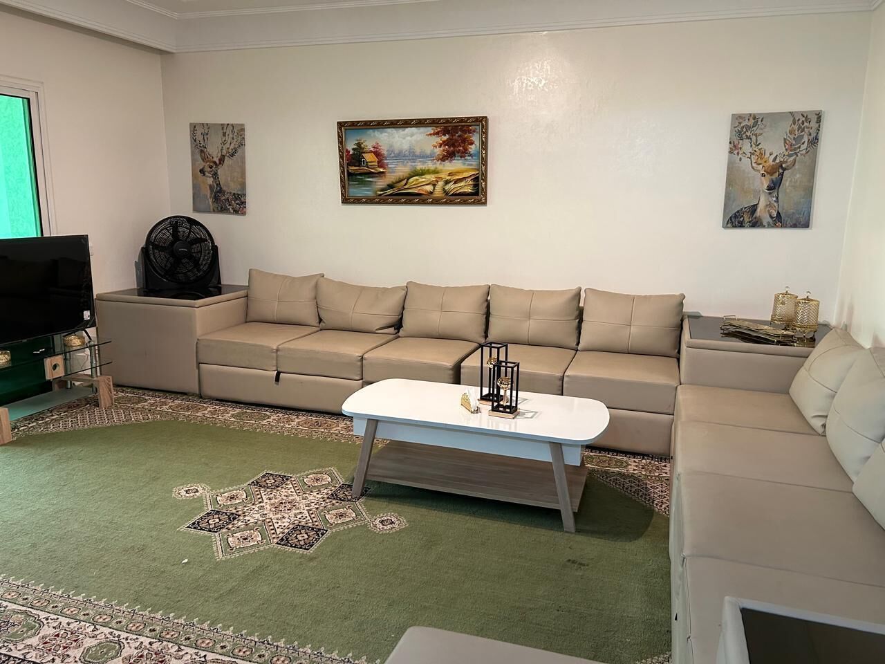 Un salon avec un canapé beige, une table basse blanche, un téléviseur et un ventilateur.
