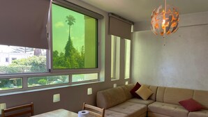 A room with a dining table, a sofa, and a large window with a view of greenery.