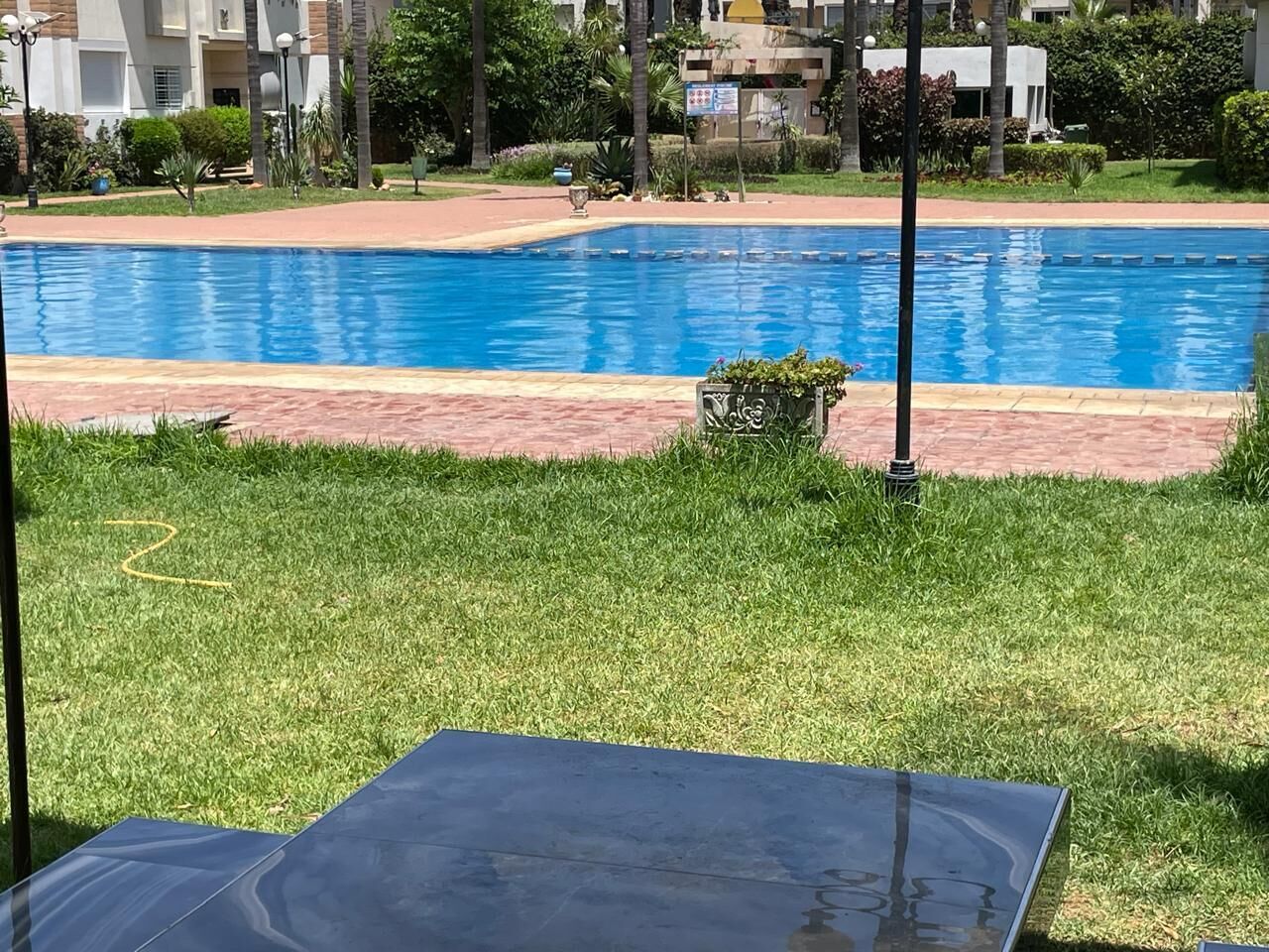 Une piscine avec une zone herbeuse au premier plan, un chemin pavé et des bâtiments en arrière-plan.