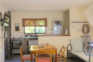 Une cuisine compacte équipée d’une cuisinière, d’un four, d’un four à micro-ondes et d’un évier. On y trouve une table à manger avec des chaises et un petit canapé avec un ventilateur.
