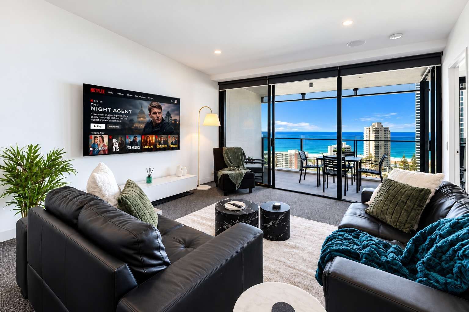 A modern living room with a flat-screen TV displaying a Netflix series, a balcony with a view of the ocean, and a seating area with leather sofas and a coffee table.